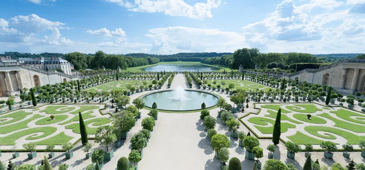 a photo of a formal French garden with topiaries, a fountain, and a viewing pool off in the distance. The sky is blue with some clouds.