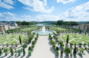 a photo of a formal French garden with topiaries, a fountain, and a viewing pool off in the distance. The sky is blue with some clouds.